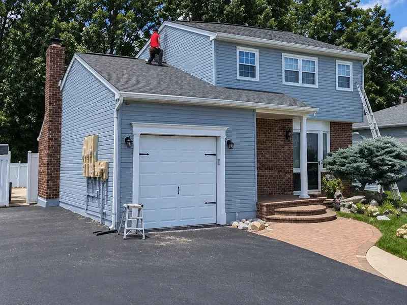 Two-story home with light blue siding, white trim, and brick chimney, painter working on the roof line